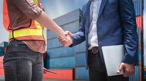 Logistics partnership: handshake between warehouse worker and business professional in front of shipping containers